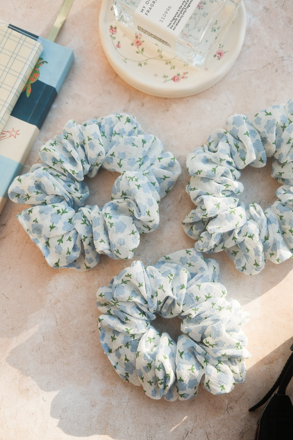 Set of floral scrunchies on a light surface with a book and container in the background.
