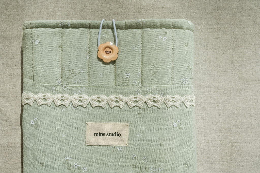Sage booksleeve with lace trim and a button, featuring 'mins studio' label, on a textured beige background.