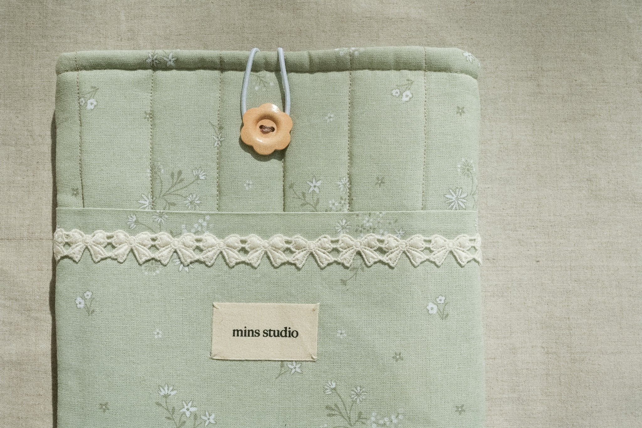 Sage booksleeve with lace trim and a button, featuring 'mins studio' label, on a textured beige background.