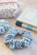 Blue scrunchie with black polka dots on a light surface with a floral pouch and notebook in the background.