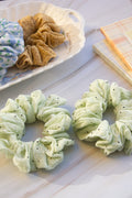 Two green scrunchies on a white surface with a basket of items in the background.