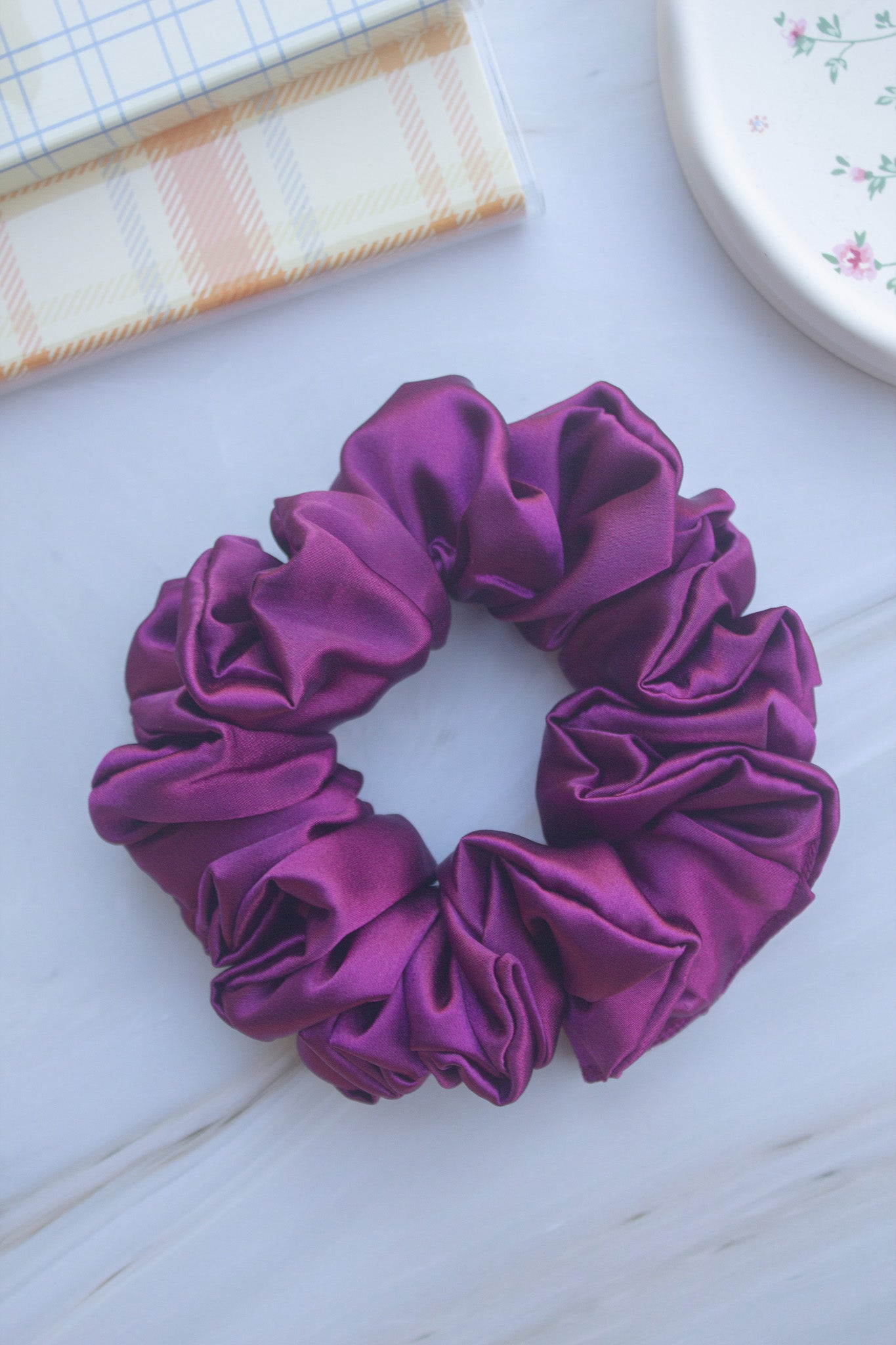 Purple scrunchie on a light surface with a checkered cloth and floral-patterned object in the background.